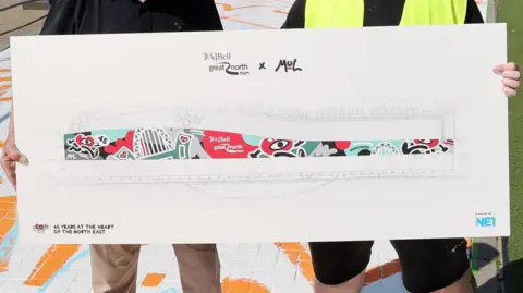 Great North Run A large piece of card showing the design being created. A cartoon heart with arms and legs is depicted running with the Tyne Bridge in the background. Two people, presumably Mul and Foster, are holding the card although only their upper torsos, hands and part of their legs can be seen. They are standing on the mural.