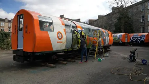 A person scrubbing graffiti from a orange and yellow subway carriage.