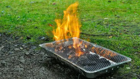 A disposable barbecue straddles a soily patch and grass as it burns.