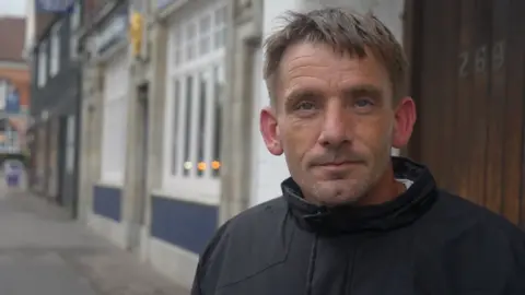 Jamie Niblock/BBC Raymond Burgess stands on a street with a blue and white building behind him. He wears a black jacket with a white T-shirt underneath