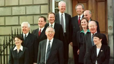 PA Media A group of 11 people politicians in suits standing in front of the front door of Bute House. Donald Dewar is front and centre, Jim Wallace is standing at the back next to Henry McLeish. They all look very happy. 