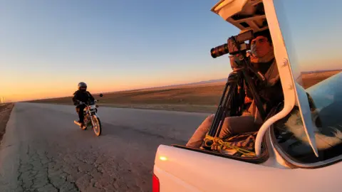 Gandom Films A man films a motorcyclist from a white pick-up truck. He is holding a camera, with the motorcyclist on the open road behind him. The backdrop is the orange glow of a sunset, set against a light blue twilight sky, on flat barren terrain.