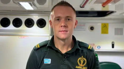 Stephen Doherty in a green ambulance uniform looking down the camera. He has short cropped, fair hair and is standing in the back of an ambulance dispatch van.