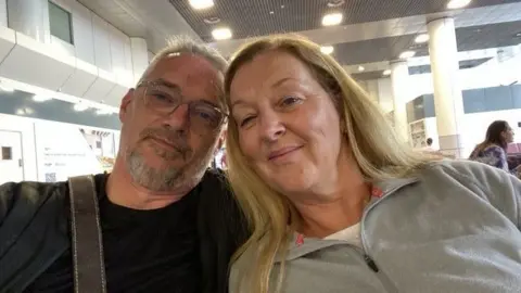 Family photopgraph A man and woman in an airport. Gary, on the left, has grey hair and a beard and is wearing spectacles. Julie, on the right, has long blond hair and is wearing a grey top.