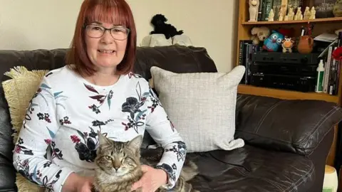 Clare Frisby/BBC A woman sits on a sofa wearing a floral white shirt holding a tabby cat with one eye