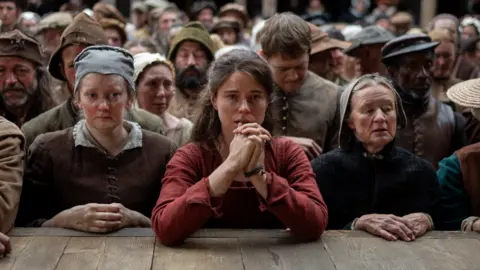 2025 FOCUS FEATURES LLC Jessie Buckley starring as Agnes Shakespeare in Hamnet. She is wearing a red dress and has her elbows on a wooden stage. She is looking out towards the stage and is standing among a crowd of people.