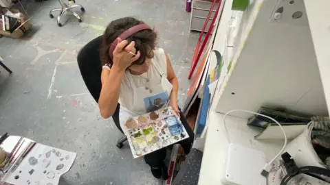 Riki Auton A over head shot of a woman with brown curly hair wearing a pink headband, she has on a white apron and is holding a palette with different colors of paint. 
