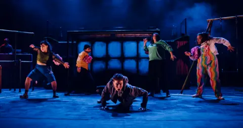 National Theatre Wales Five performers captured under dark blue stage lights. The centre performer is crawling across the floor and stares directly at the camera while other characters pose in abstract positions around them. 