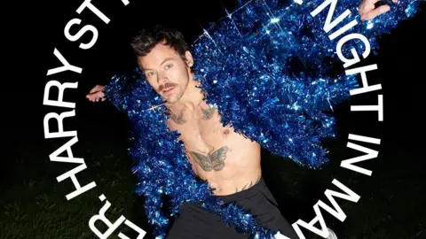 Harry styles, who is wearing grey trousers and a a blue jacket made out of tinsel, is staring at the camera in this promotional photograph. 