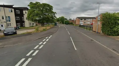 Google A Google street view image of a residential road. A junction is on the left and behind that is a cream and black coloured block of flats, with a car parked in the car park in front of it and a large tree. On the right are other residential buildings, fences and a bush.