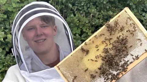 A teenage boy wearing a beekeeping suit and blue gloves holds a frame full of bees up at the camera.