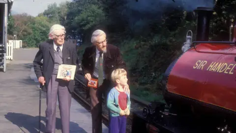 Talyllyn Railway collection Three people standing on a platform next to a red steam engine. On the left is Reverend Wilbert Awdry, who wrote the Thomas the Tank Engine books. He is an old man with white hair who is wearing a jacket and tie with v-neck jumper. His son stands next to him, a slightly younger man with a beard, also wearing a jacket and tie. The third person is Rev Awdry's grandson, a child who's looking at the steam locomotive. The adults are carrying copies of the Thomas the Tank Engine books, while Rev Awdry's son puts a protective hand on the grandson's shoulder.
