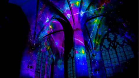 Luxmuralis Artistic Collaboration/Peter Walker/Carrie Potter A cathedral ceiling and arched window lit up in blue, pink, purple and green lights