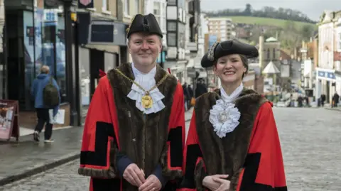 A man and a woman on a cobbled high street wearing red and brown robes and historical white collars. They are also wearing traditional black hats and mayoral medallions.