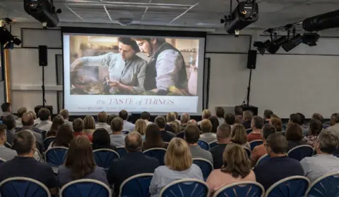 Stephen Evans Seven rows of people sat on blue chairs watching a screen with a film projected onto it.
