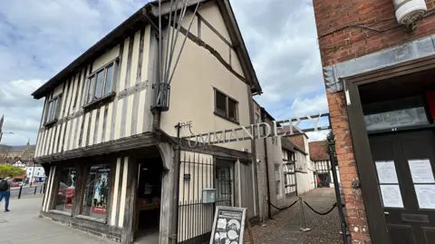 This is an image of The Commandery, a museum in Worcester. It is an old, white building with black beams on the front of the building. There is an alleyway net to the building which has a sign that reads "The Commandery" above it.
