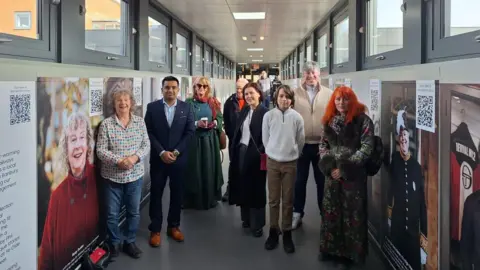 Eight residents, both men and women, stand in front of their portraits, which are mounted on the walls either side of the overbridge at Banbury Station. 