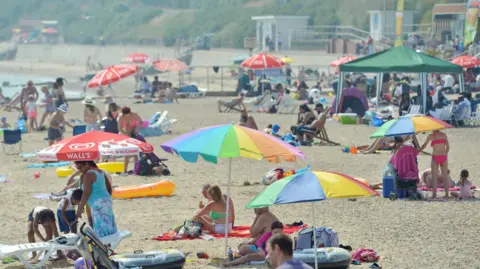 PA Media People enjoying the sunshine on the beach in Clacton-on-Sea. There are people sitting beneath colourful umbrellas, lying on towels and walking around the sand.