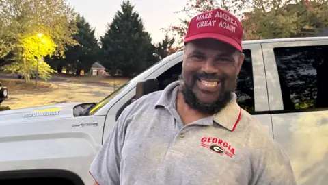 Gerald Cambor poses for a photo in front of a car wearing a "make America great again" red hat