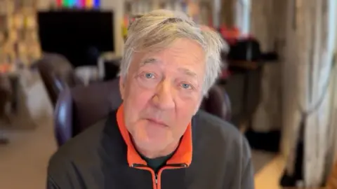 Spark UK Stephen Fry - a man looking up into his web camera with the background blurred, but it looks like a study.