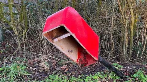 A view of a damage postbox. The pole that the box is on is at an angle and the front door of the postbox is missing.