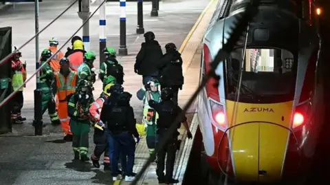 Getty Images A yellow and red and white train parked on the station, with emergency workers standing next to it on the platform.