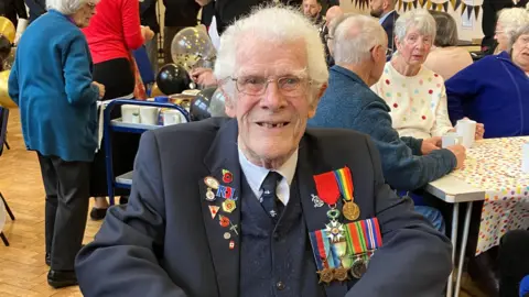 Jim Grant is wearing a dark blue jacket with medals pinned to it and is sitting in a room surrounded by friends celebrating his 100th birthday 