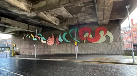 BBC A view of Traffic Flow on the side of a concrete wall. It is a sculpture made of stainless steel painted in a variety of colours including red and green formed of squiggles and wiggly lines.