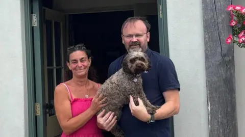 Elm Tree Inn A woman with brown hair wearing a pink dress holding a grey dog. A man in a blue top and camel coloured trousers also holding the dog.