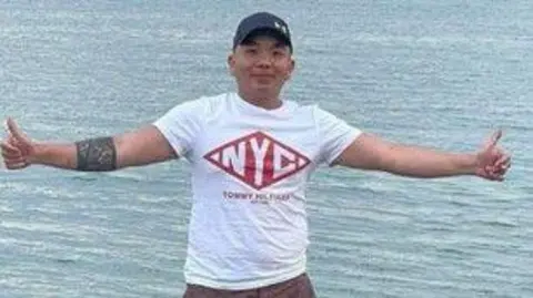 Vo Thai Son is standing in front of a body of water with his arms stretched out to his side and his thumbs up. He is wearing a white top with 'NYC' written in the centre. He has a distinct band tattoo on his forearm. He is smiling at the camera and also wears a blue baseball cap.