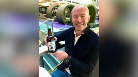 Contributed Alan Shearer is pictured holding a bottle of Away Days beer. He is smiling at the camera. He is bald and wearing a navy cardigan with a shirt on underneath. 