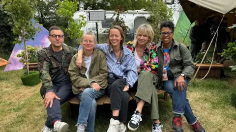 (Left to Right) - Jonathan Rothery, Alison Howe, Emily Eavis, Zoe Ball, Lorna Clarke all sat on a bench backstage at Glastonbury Festival.
