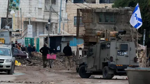 Reuters Palestinians walk past an Israeli military vehicle and an armoured bulldozer during a major operation by Israeli security forces in Jenin, in the occupied West Bank (22 January 2024)