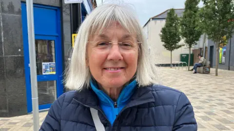 A woman with grey, shoulder-length hair and clear glasses stood in a street. She is wearing a dark blue jacket and looking at the camera