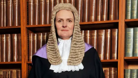 Avalon Judge Alison Raeside looks straight at the camera. She is wearing a wig and gown and is pictured in front of a bookshelf
