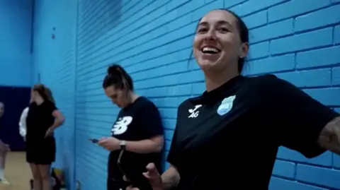 Dorset Futsal Club and K1DProductions Amie Gray wearing a black sports kit and smiling. She is standing against a blue brick wall.