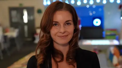 A head-and-shoulders picture of Rieke Dickinson. She has long brown hair in loose bouncy curls and blue eyes. She is wearing a black top and smiling at the camera. The background is blurred.  