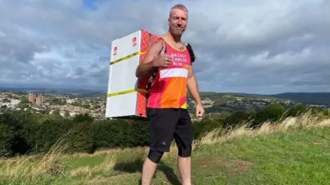 Joanne Graham A man with closely cropped grey hair and a pink running vest stands on a hillside with a large fridge strapped to his back