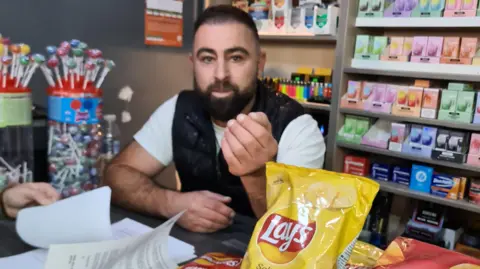 A man with short dark hair and a beard standing behind a shop counter. There are various items on display, including a large packet of crisps and lollipops.