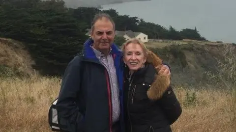 Terry, on the left, is wearing a patterned shirt and a black coat and has short brown hair. Wendy J. Tomlin-Hess is on the right and has short dark blonde hair, and is wearing a black coat. They are stood on a hill. 