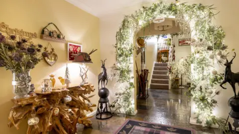 Kirsty Anderson Festively decorated hallway with a rustic wooden console table, Christmas ornaments, and a large archway adorned with greenery, white flowers, and twinkling lights leading to a staircase