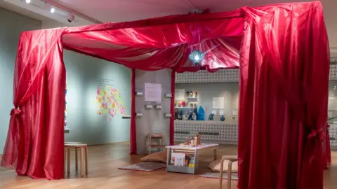St Albans Museums A museum or exhibition space featuring a vibrant red canopy structure made of sheer fabric, creating an enclosed area. Inside the canopy, there is a low white table with books and decorative items, surrounded by floor cushions and small stools.