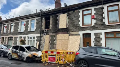 A stone terraced house with fire damage around the windows and doors, and wooden boards covering them. In front are two cars parked, one is badly fire damaged. 