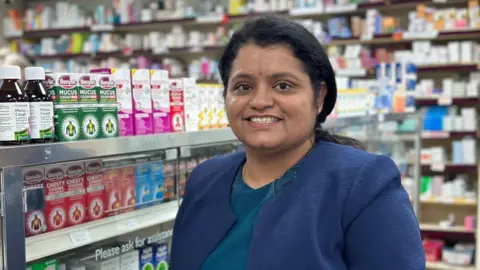 Sri is pictured in her pharmacy in Barmston, standing in front of shelves full of cold and flu medicine. She has long black hair which is tied back from her face. She is wearing a smart blue jacket. 