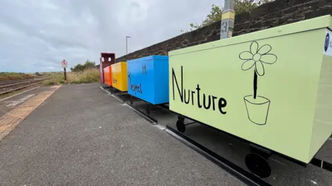 A view of a train sculpture focused on the green back carriage which has 'Nurture' and a pen drawing of a flower in a pot drawn on it. The next carriage up is blue and had 'Respect' written on it. The carriages have been placed next to fake tracks. The sculpture is on a platform next to a real railway line.