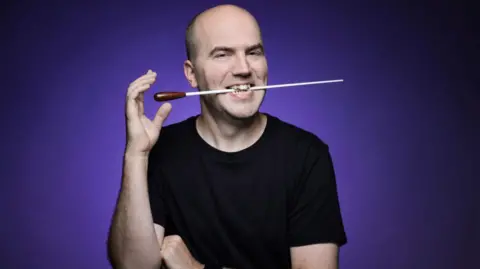 Sam Watts Composer Sam Watts in front of a purple background, wearing a black T-shirt