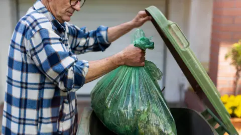 A man wearing a blue and white fleece puts a green bag of rubbish into a bin.