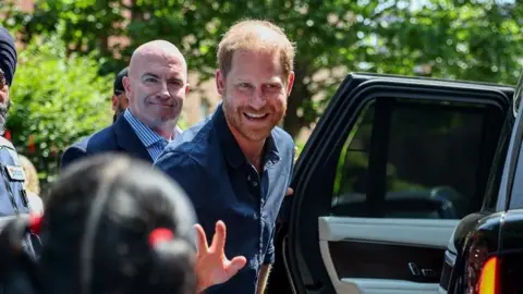 Reuters Britain's Prince Harry waves at a child and a crowd as he gets into his car after leaving a hospital visit. He is wearing a blue shirt, and is looking to the left of the camera and smiling