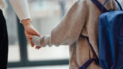 A view from behind a child and adult, holding hands. The child wears a grey cardigan and a blue rucksack. The adult has black trousers and a long-sleeved white top.
