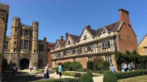 Coughton Court, a National Trust property with a castle-like main entrance. It is a Tudor house, surrounded by gardens. There are a number of visitors in the photo, viewing the property.